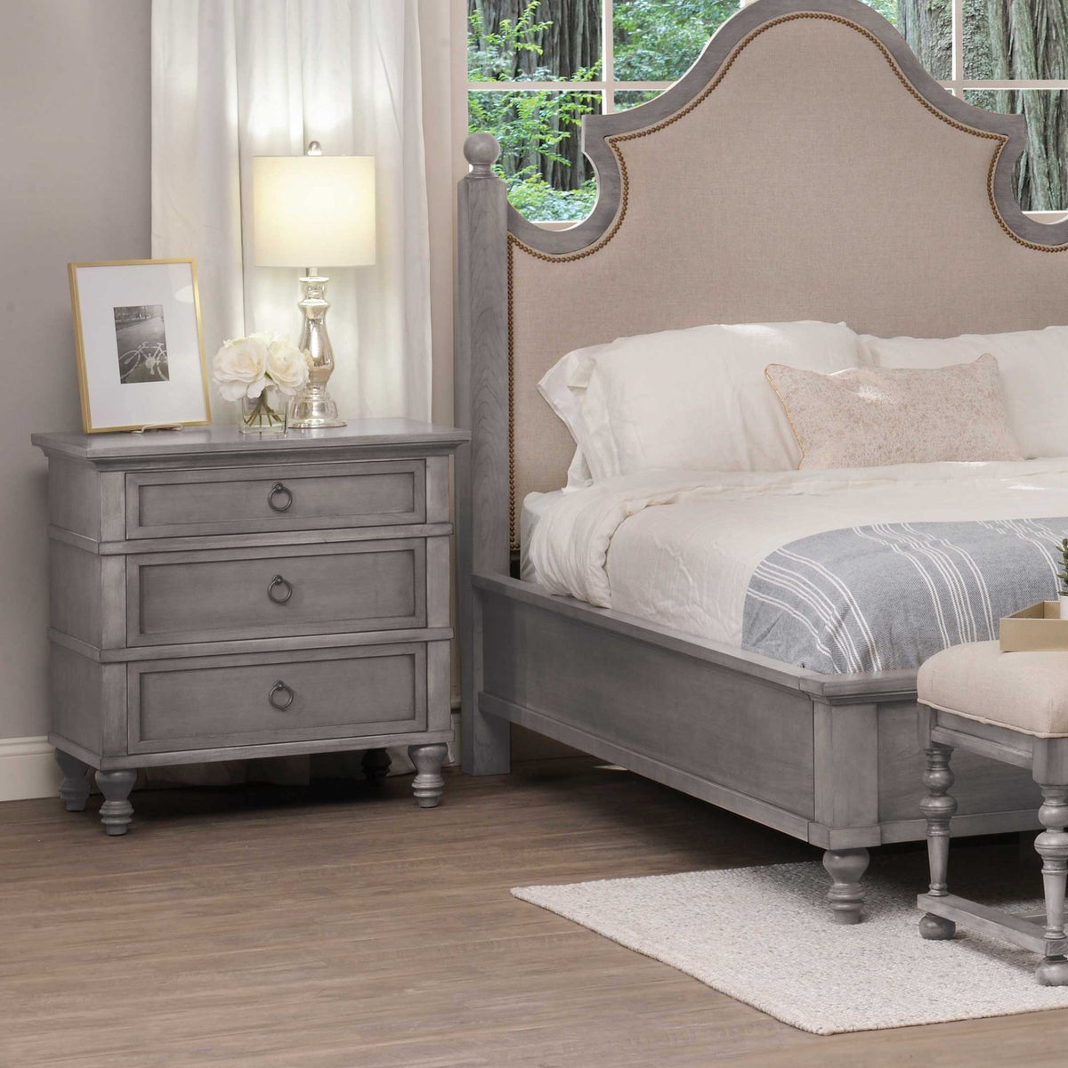 Driftwood gray nightstand with three ring-pull drawers styled next to a nailhead upholstered bed and lamp