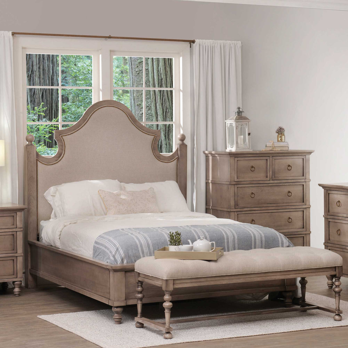 Taupe upholstered bed with nailhead-trimmed arched headboard styled with a tufted bench, chest and nightstand
