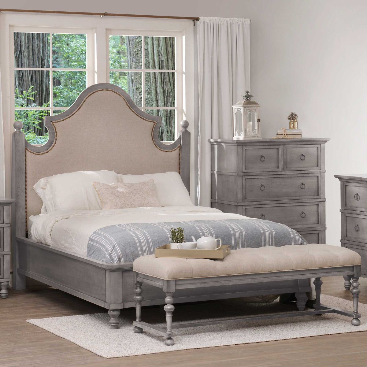 Gray upholstered bed with arched linen nailhead headboard, ball finials and tufted bench in a sunlit bedroom