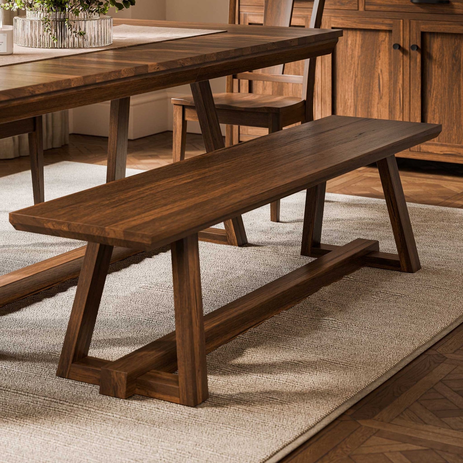 Long walnut bench with trestle legs and stretcher beside a matching dining table on a herringbone wood floor