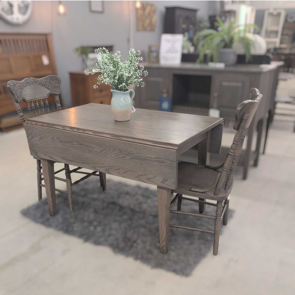 Gray stained drop leaf dining table with one leaf down and two carved spindle chairs on a furniture showroom floor