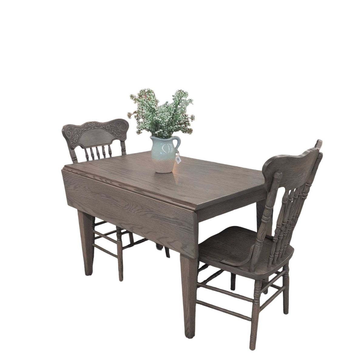 Two carved spindle back chairs at a gray washed oak drop leaf table with one leaf down and a pitcher of blossoms