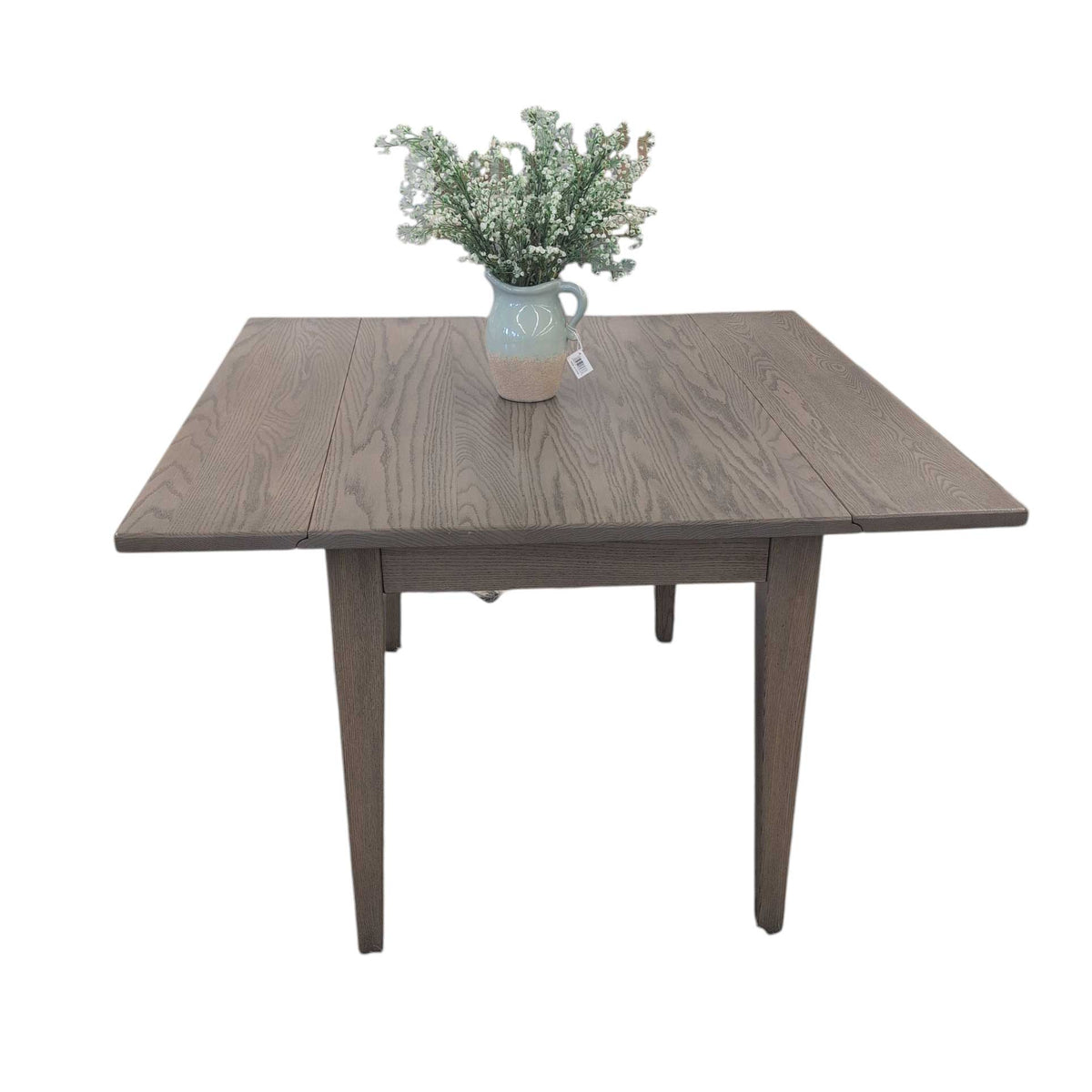 Weathered gray oak drop leaf dining table with both leaves up, tapered legs and a stoneware pitcher of blossoms