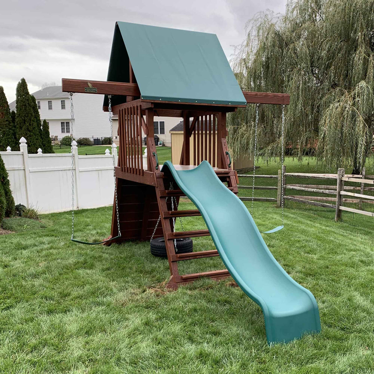 Wooden backyard playset with green tarp roof, wavy slide, tire swing and two belt swings on a fenced lawn