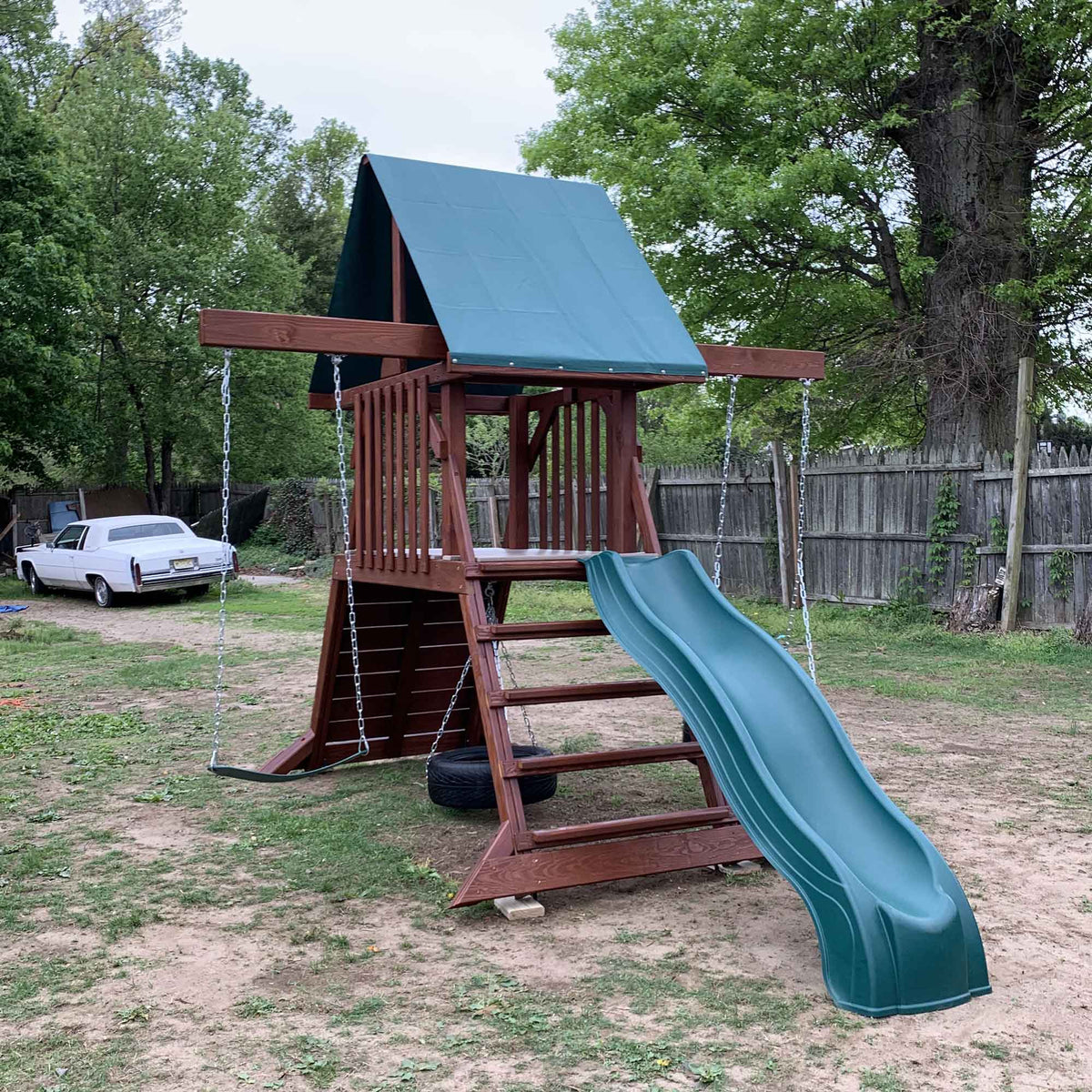 Redwood stained wood playset with green wave slide, tire swing and swing beam standing in a grassy backyard