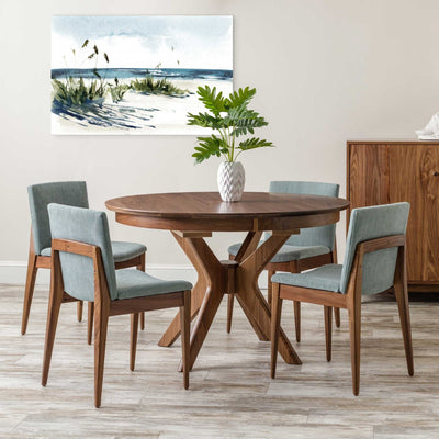 Round walnut dining table with a splayed cross pedestal base and four seafoam upholstered chairs beneath coastal artwork