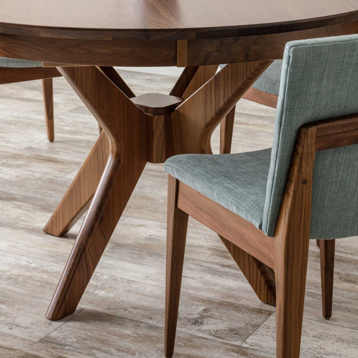 Close-up of the walnut table's crossed pedestal base joint with a seafoam upholstered dining chair alongside