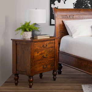 Three drawer rustic cherry nightstand with bronze bail pulls and turned legs beside a sleigh bed