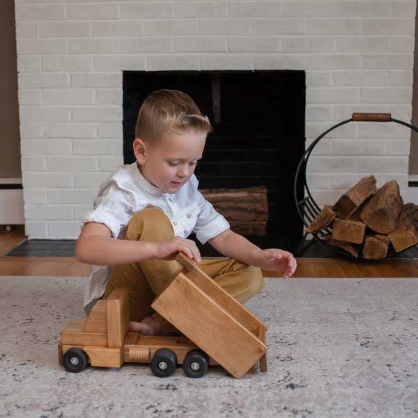 Boy in a white shirt kneeling on a rug tipping the bed of a wooden toy dump truck by a brick fireplace