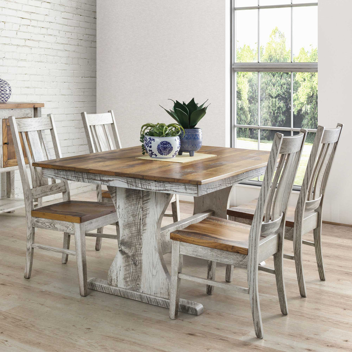 Whitewashed trestle dining table with a warm reclaimed wood top and six matching slat back chairs in a bright brick walled room