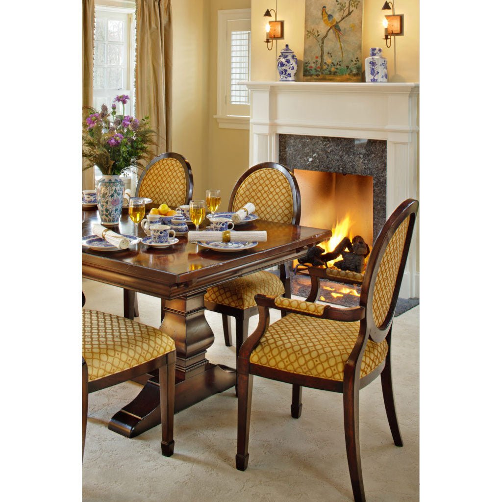 Cherry pedestal dining table set for a meal with gold patterned oval back chairs beside a lit fireplace