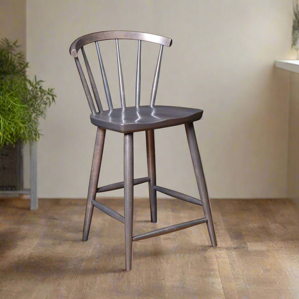 An Amish Tussock Windsor Bar Stool with a dark finish, featuring a rounded seat and spindle back design, placed in a room with a wooden floor and a plant in the background.