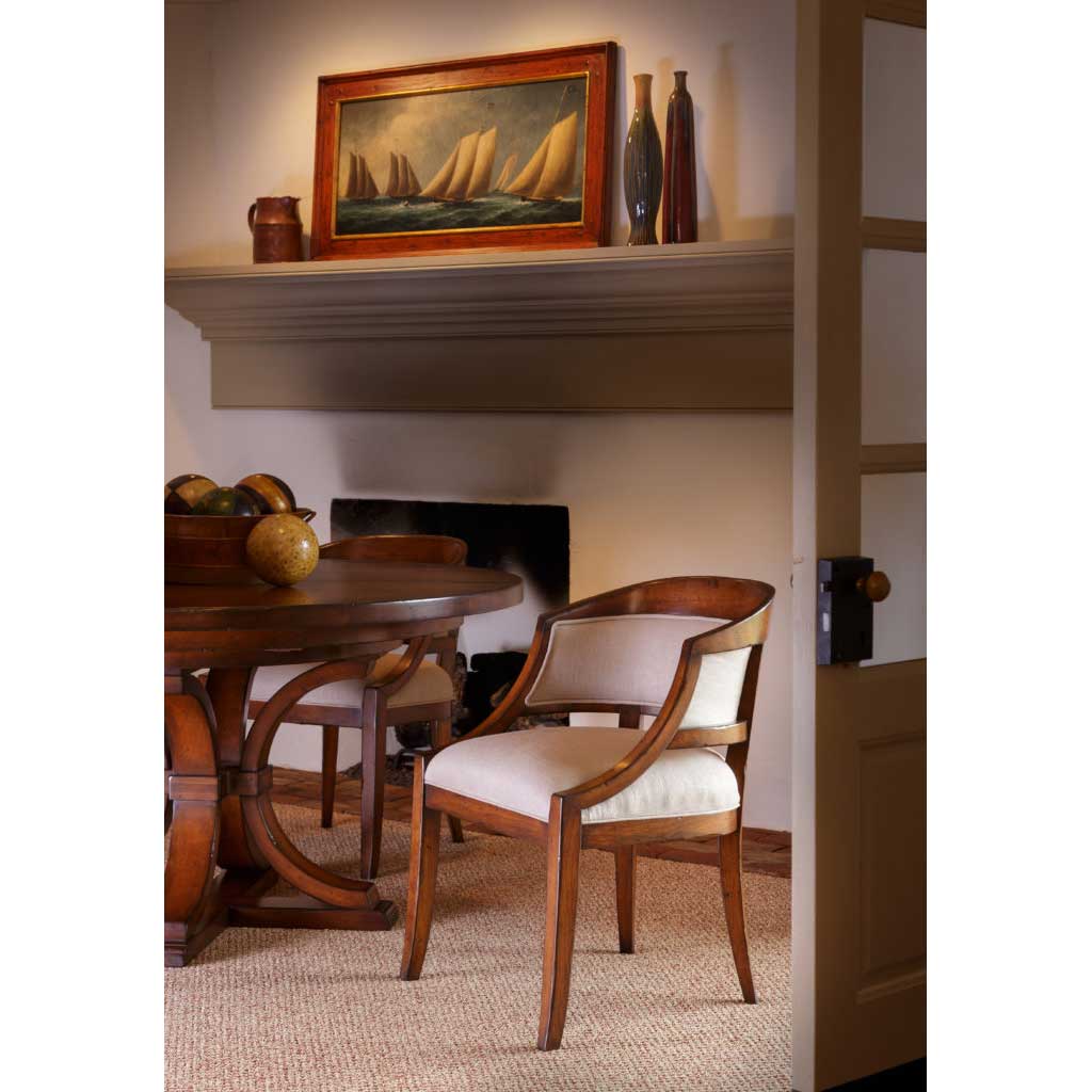 Cherry barrel back arm chair with cream linen upholstery beside a round pedestal table near a fireplace mantel