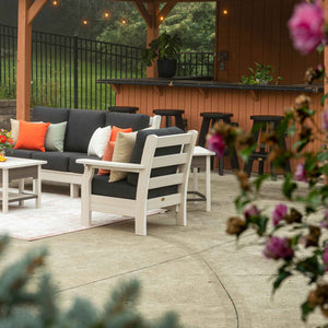 White poly patio chair and sofa with dark gray cushions and orange pillows arranged near a backyard bar pavilion