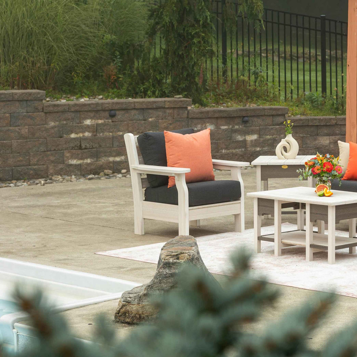 White poly patio chair with charcoal cushions and orange pillows next to matching side and coffee tables by a pool