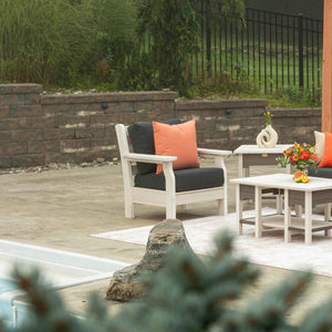 White poly patio chair with charcoal cushions and orange pillows next to matching side and coffee tables by a pool