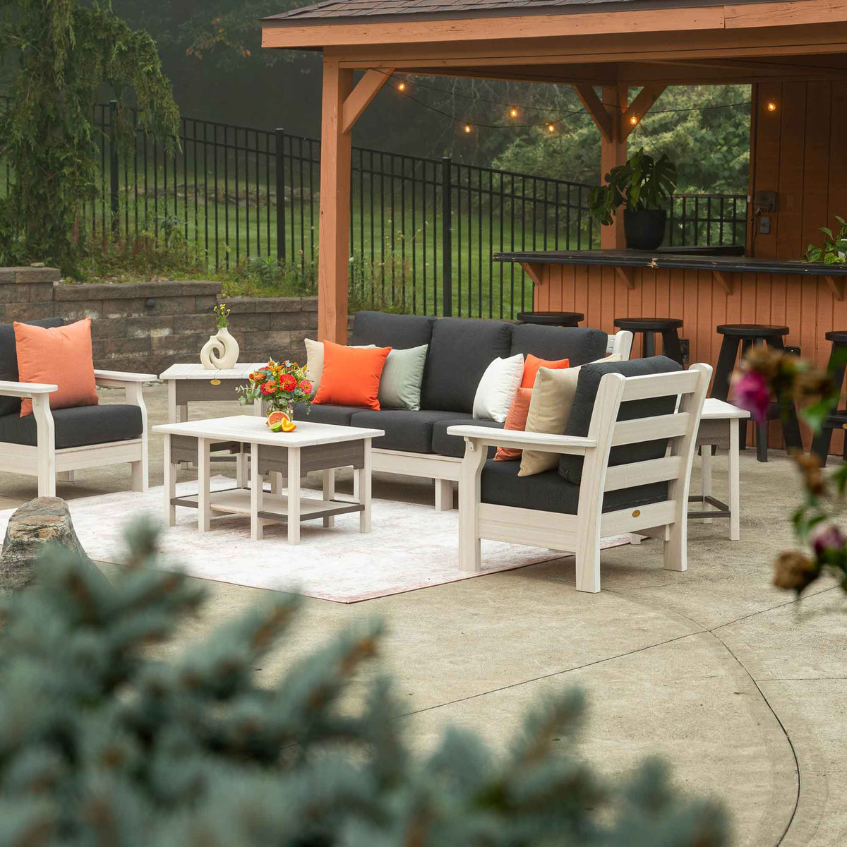 White poly sofa and chairs with charcoal cushions and orange pillows around a coffee table near a backyard bar