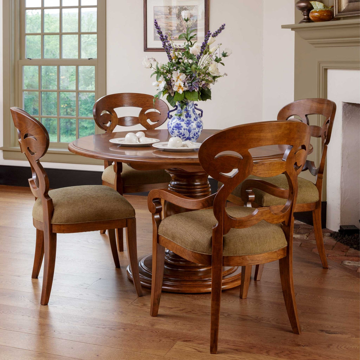 Round cherry pedestal dining table with four scrolled open back chairs and olive cushions in a bright dining room
