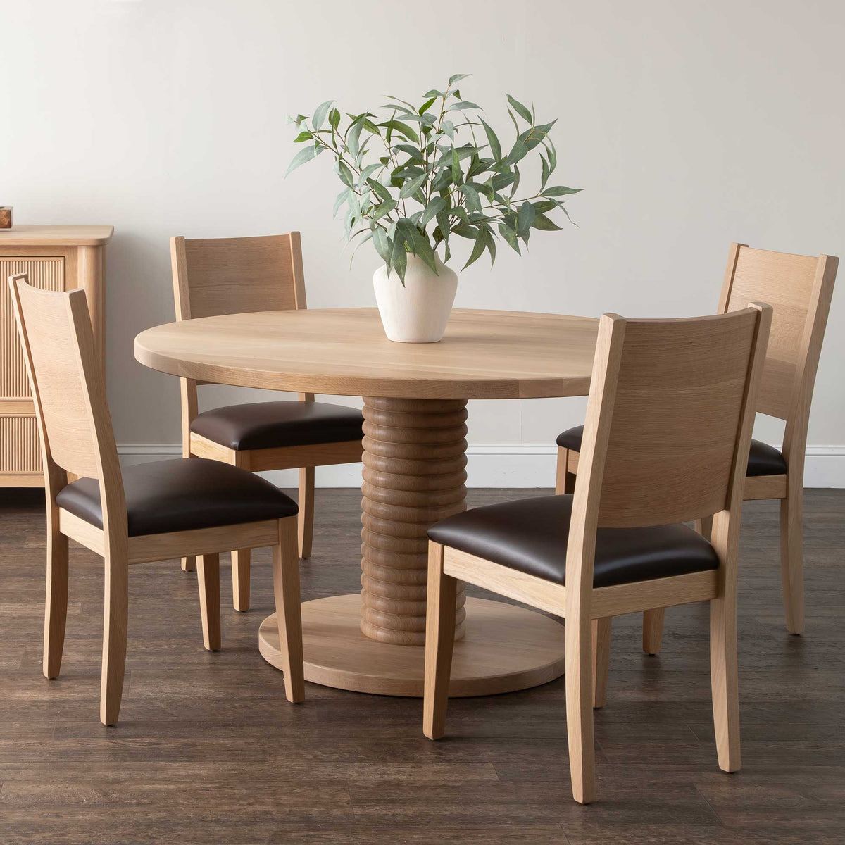 Round white oak dining table with a ribbed column pedestal and four chairs with dark leather seats in a modern room