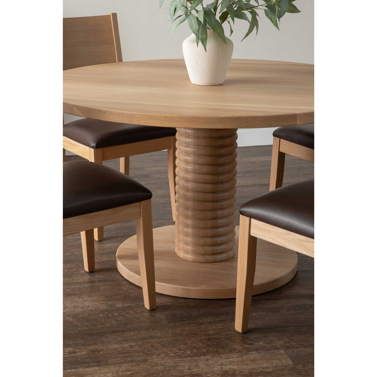White oak round dining table with a ribbed pedestal base, leather seat chairs and a white vase of eucalyptus
