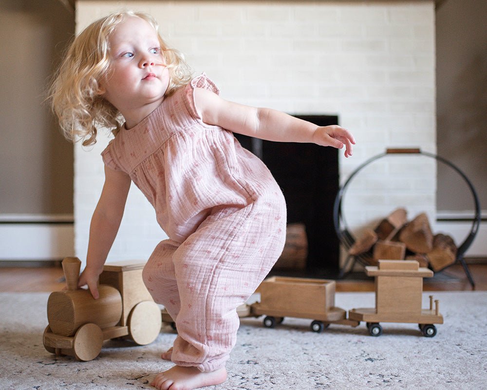Toddler in a pink romper pushing a wooden toy train with engine and cargo cars across a living room rug