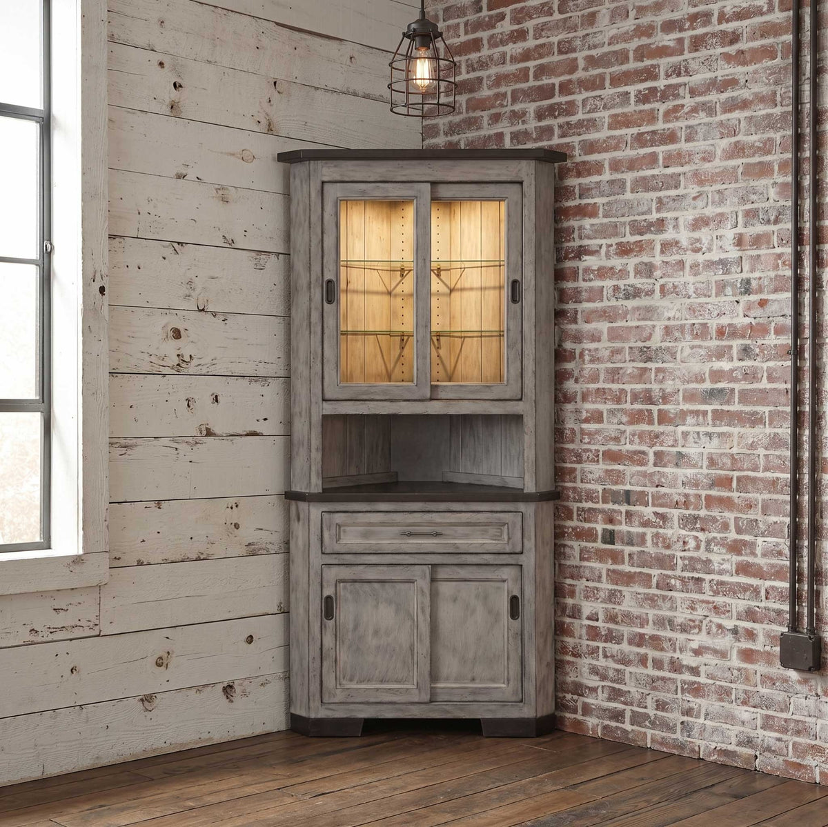 Weathered gray 31 inch corner hutch with lit sliding glass doors, dark top, one drawer and paneled base in a rustic corner