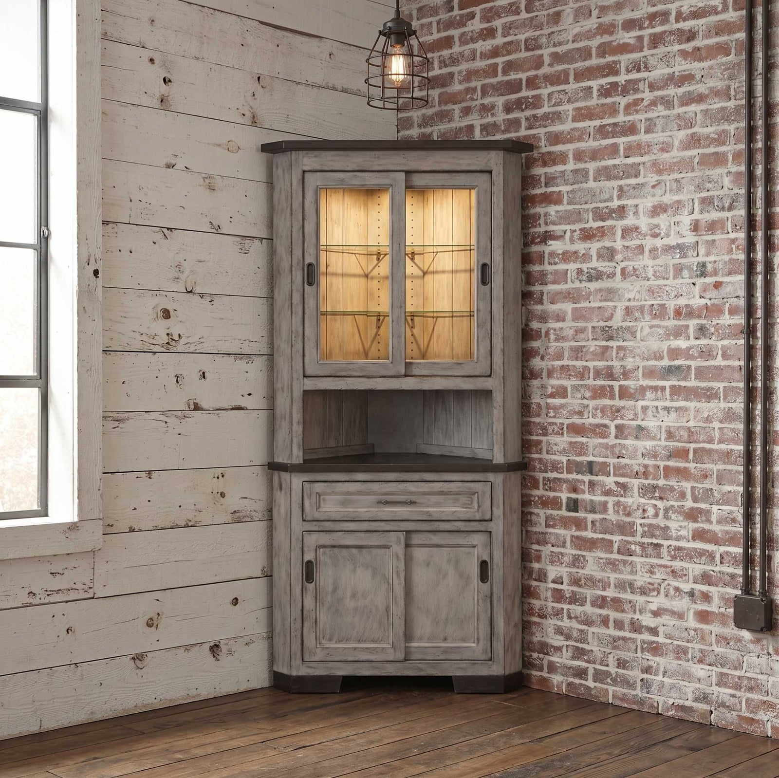 Weathered gray 31 inch corner hutch with lit sliding glass doors, dark top, one drawer and paneled base in a rustic corner