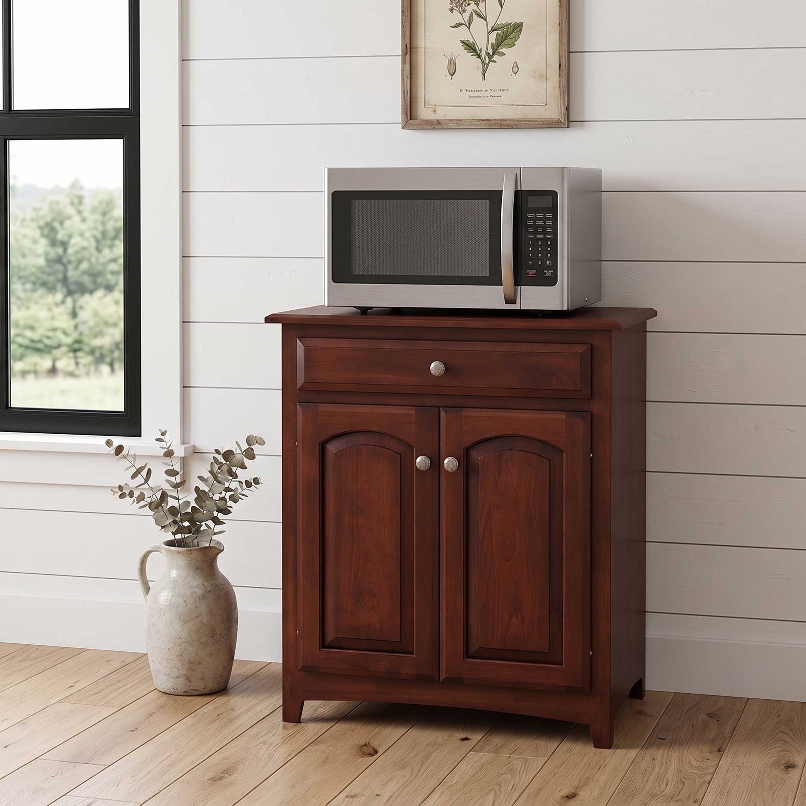 A dark brown wooden kitchen cabinet with a single drawer and two arched doors, styled with a silver microwave on top, set against a white shiplap wall with a window and a vase of eucalyptus nearby.