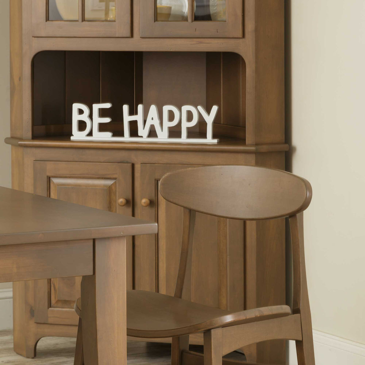 Corner hutch in warm brown beside a dining table and round back chair, with a white Be Happy sign on the shelf