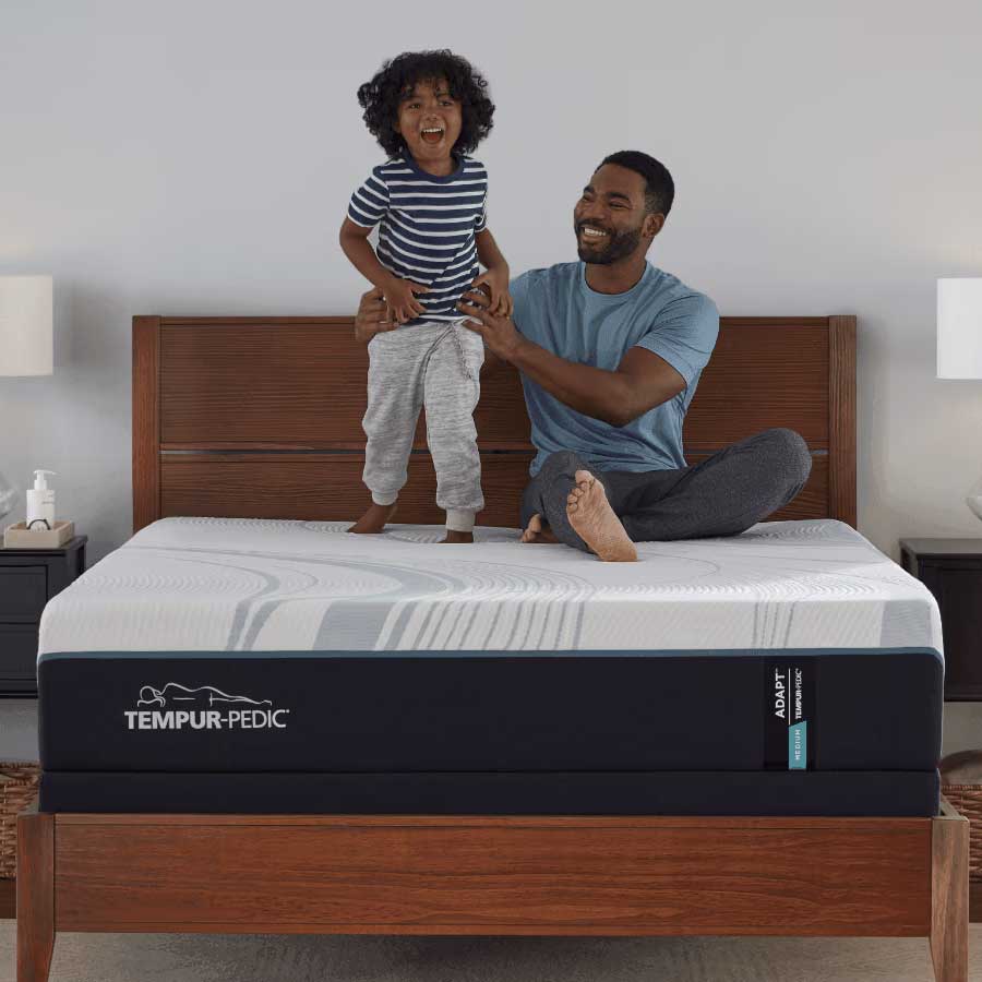 Father steadying a laughing toddler standing on a white foam mattress with navy border in a wood panel bed