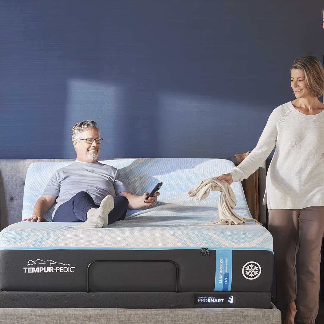 Man with a remote reclining on a cooling mattress raised on an adjustable base while a woman lays a throw down