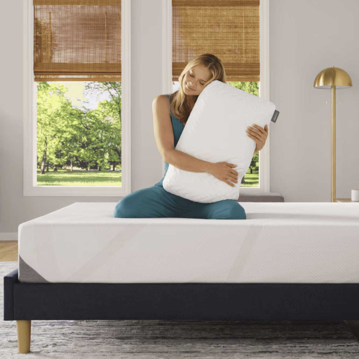 Woman kneeling on a bare mattress hugging the quilted white pillow in a bright bedroom with woven shades