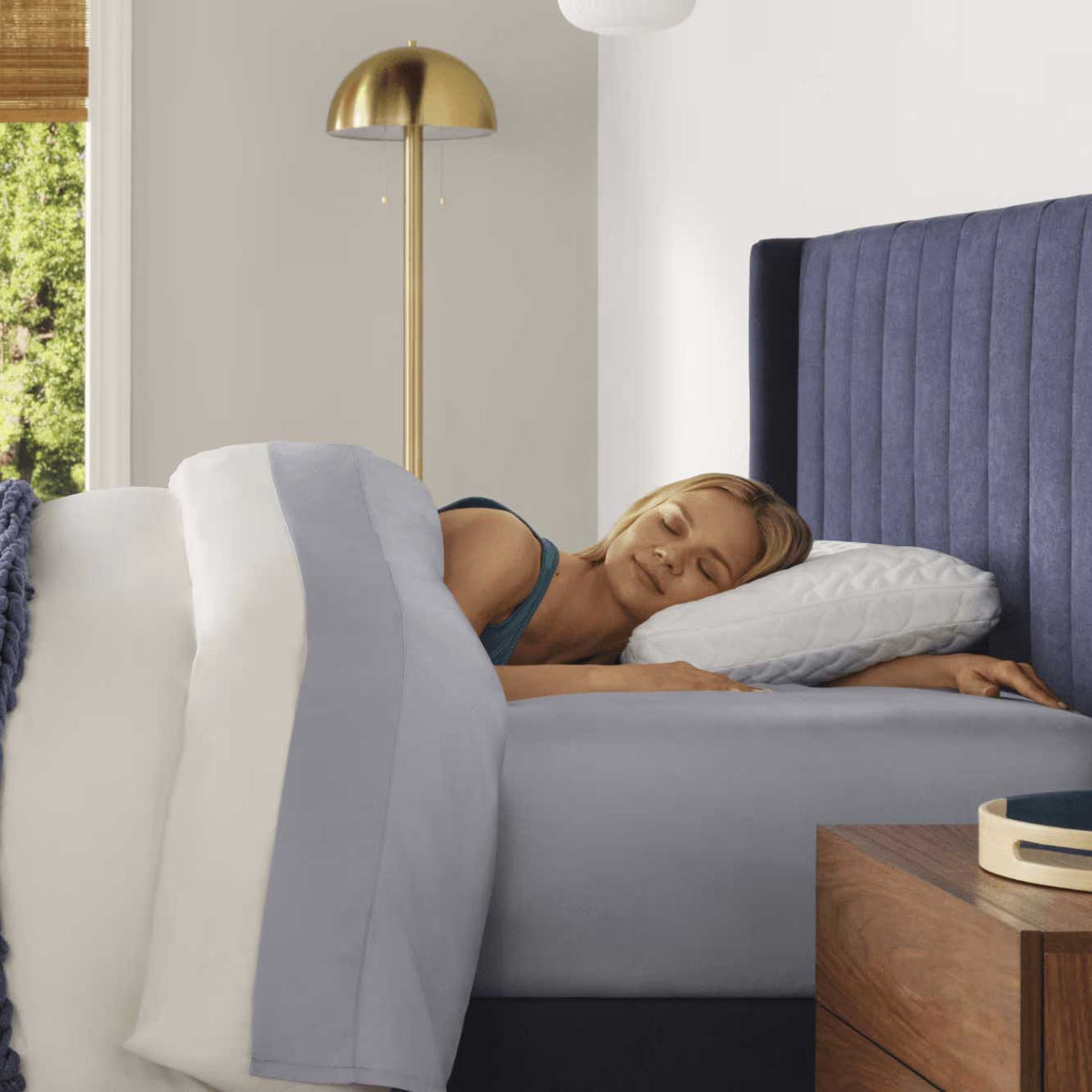 Woman sleeping on the quilted pillow in a navy upholstered bed with pale gray sheets and a brass floor lamp