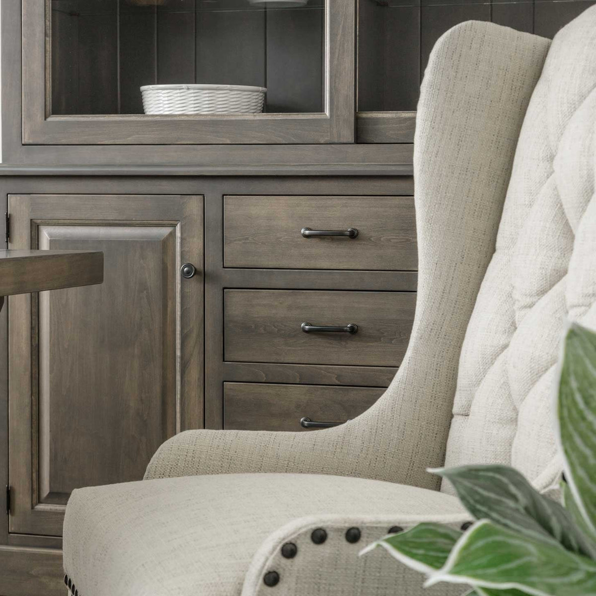 Detail of a gray sideboard with paneled door and three drawers beside a tufted linen dining chair with nailhead trim