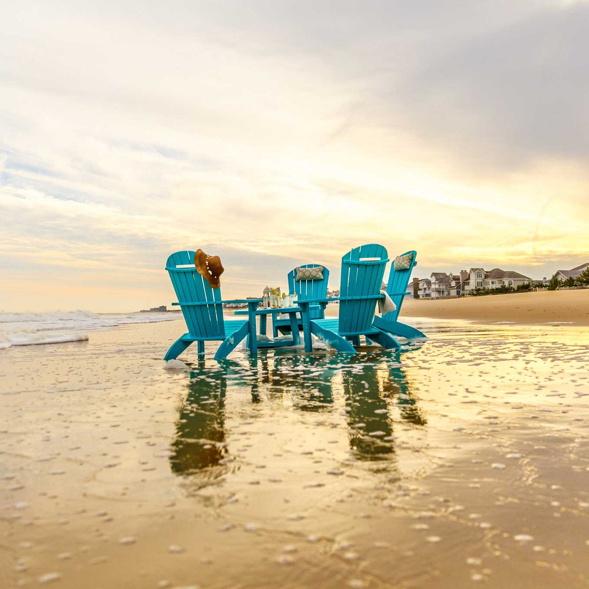 Three aqua blue poly Adirondack chairs with a side table on wet sand at sunset by the shoreline