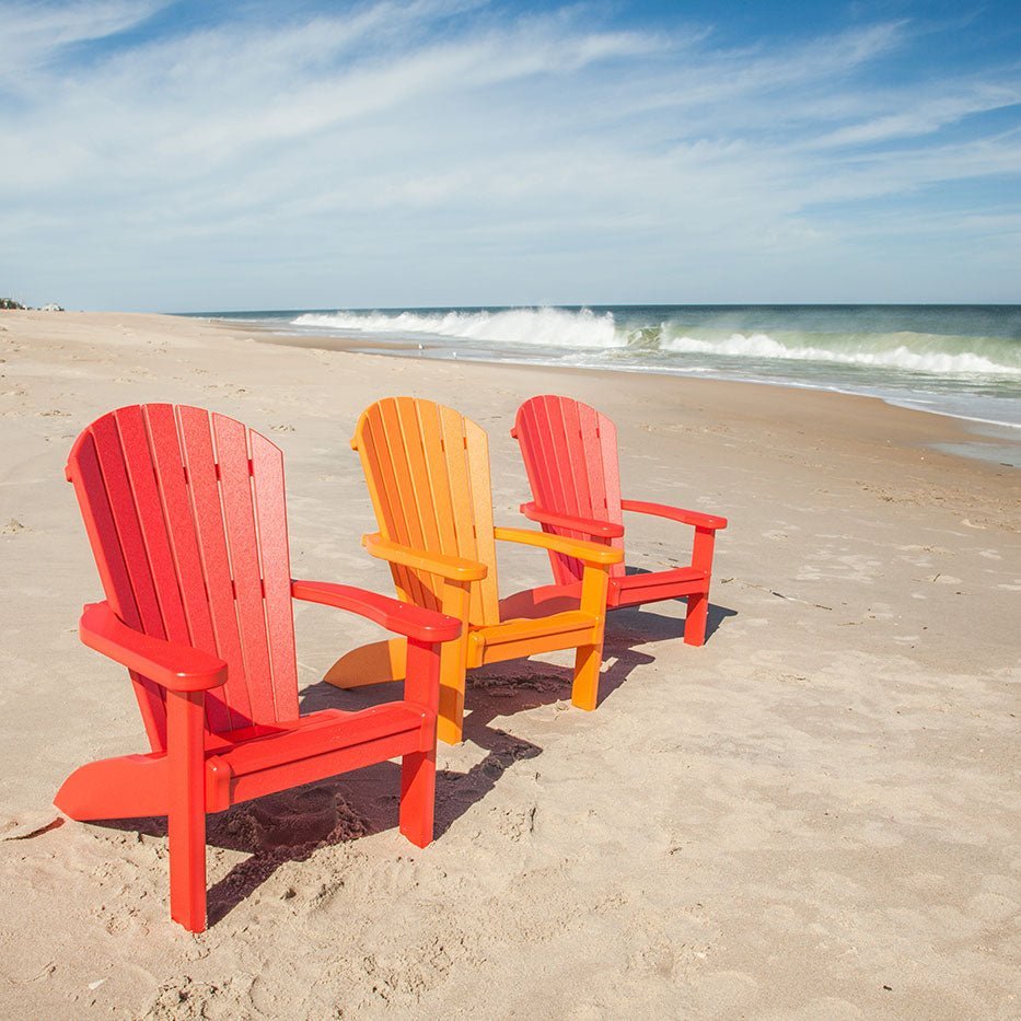 Three brightly colored red and orange poly Adirondack chairs facing the surf on an open beach