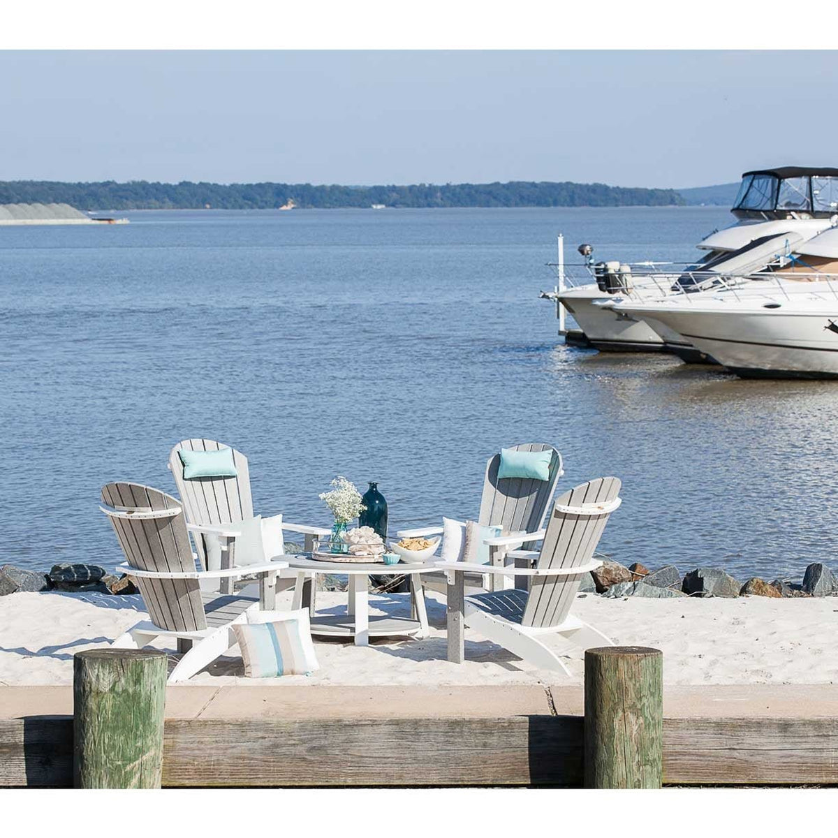 Four white and gray Adirondack chairs around a low table on a sandy dock beside moored boats
