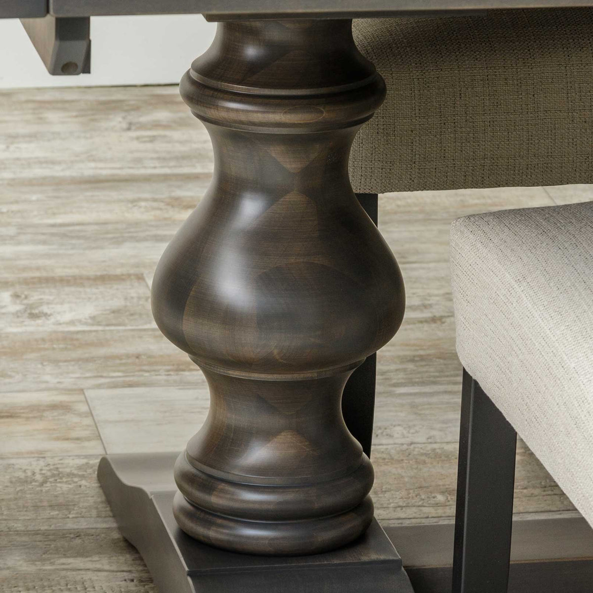Close view of the turned baluster pedestal and plank foot of a dark gray dining table between upholstered chairs