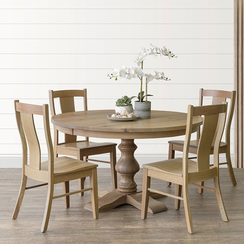 Round natural oak pedestal dining table with four light wood slat-back chairs against a white shiplap wall
