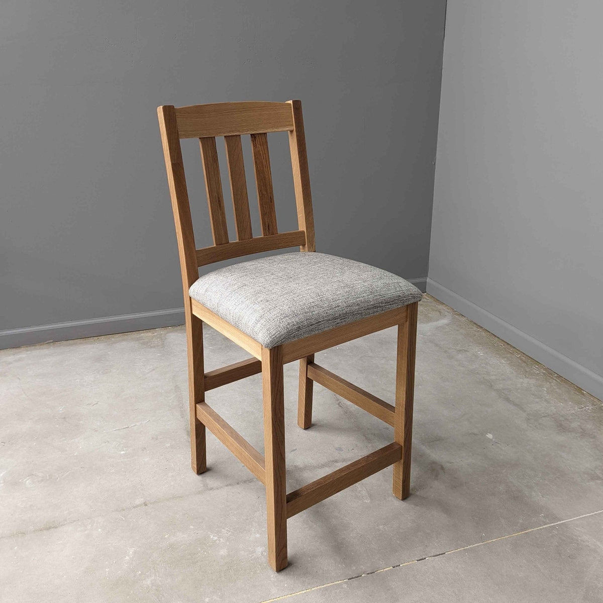 Light oak counter stool with three vertical back slats and a gray woven fabric seat in a concrete-floored room