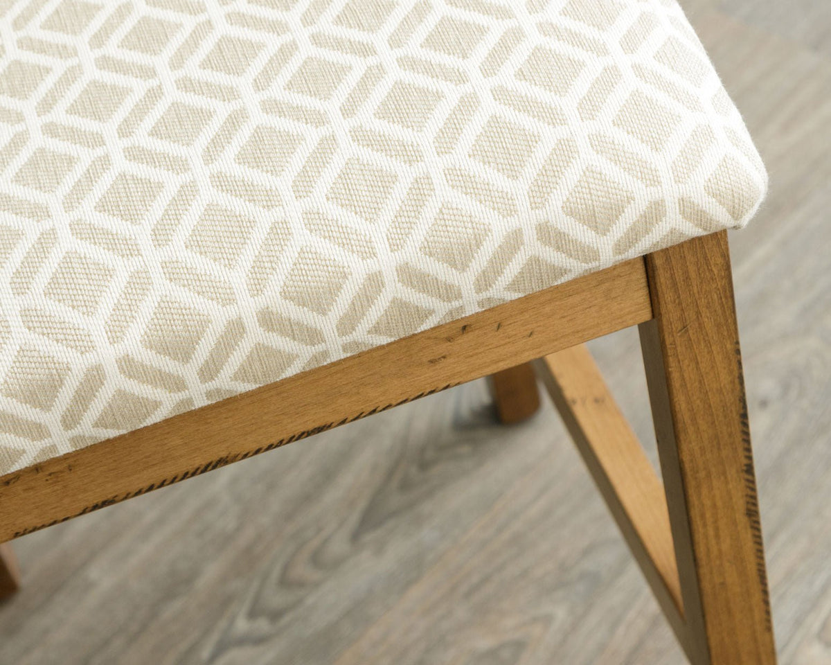 Close view of the cream geometric upholstered stool seat and distressed maple leg with box stretcher
