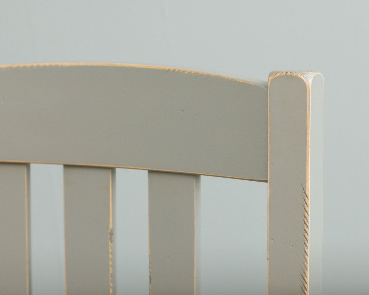 Detail of the curved top rail and slats of a gray painted dining chair with distressed edges