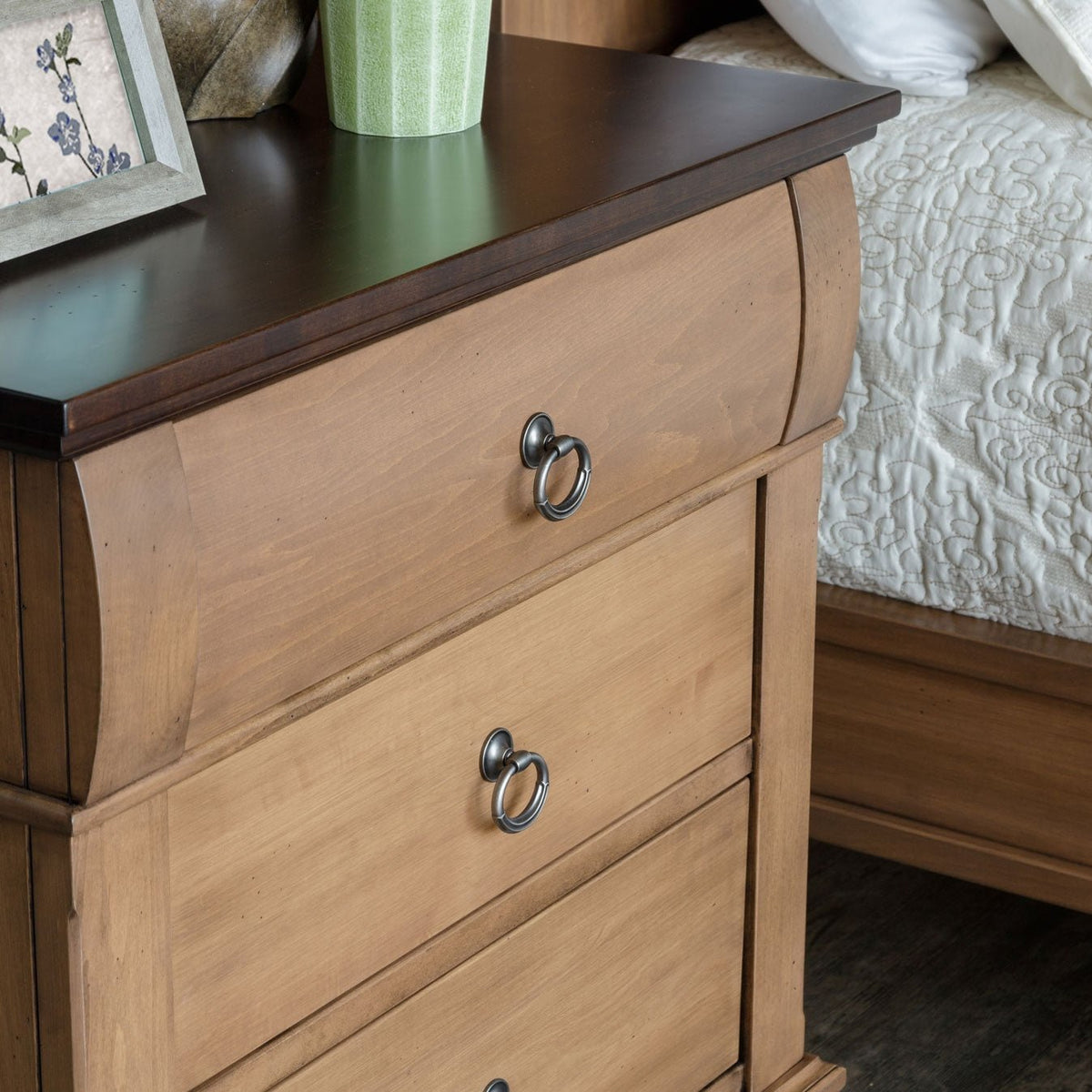 Corner detail of a light wood bedside chest with dark top holding a framed photo and green vase beside a white bed