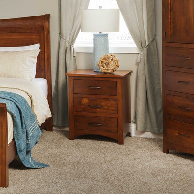 Cherry three-drawer nightstand with bar pulls beside a sleigh bed and a blue lamp in a carpeted bedroom