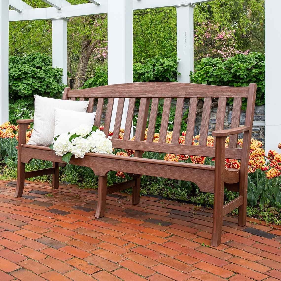 Long mahogany brown poly garden bench with an arched slat back and armrests, on a brick patio