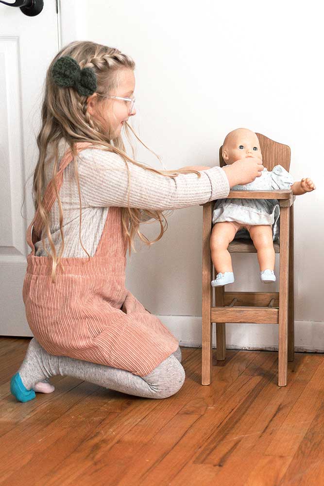 Young girl in pink overalls kneeling on a hardwood floor feeding a baby doll seated in a wooden doll highchair