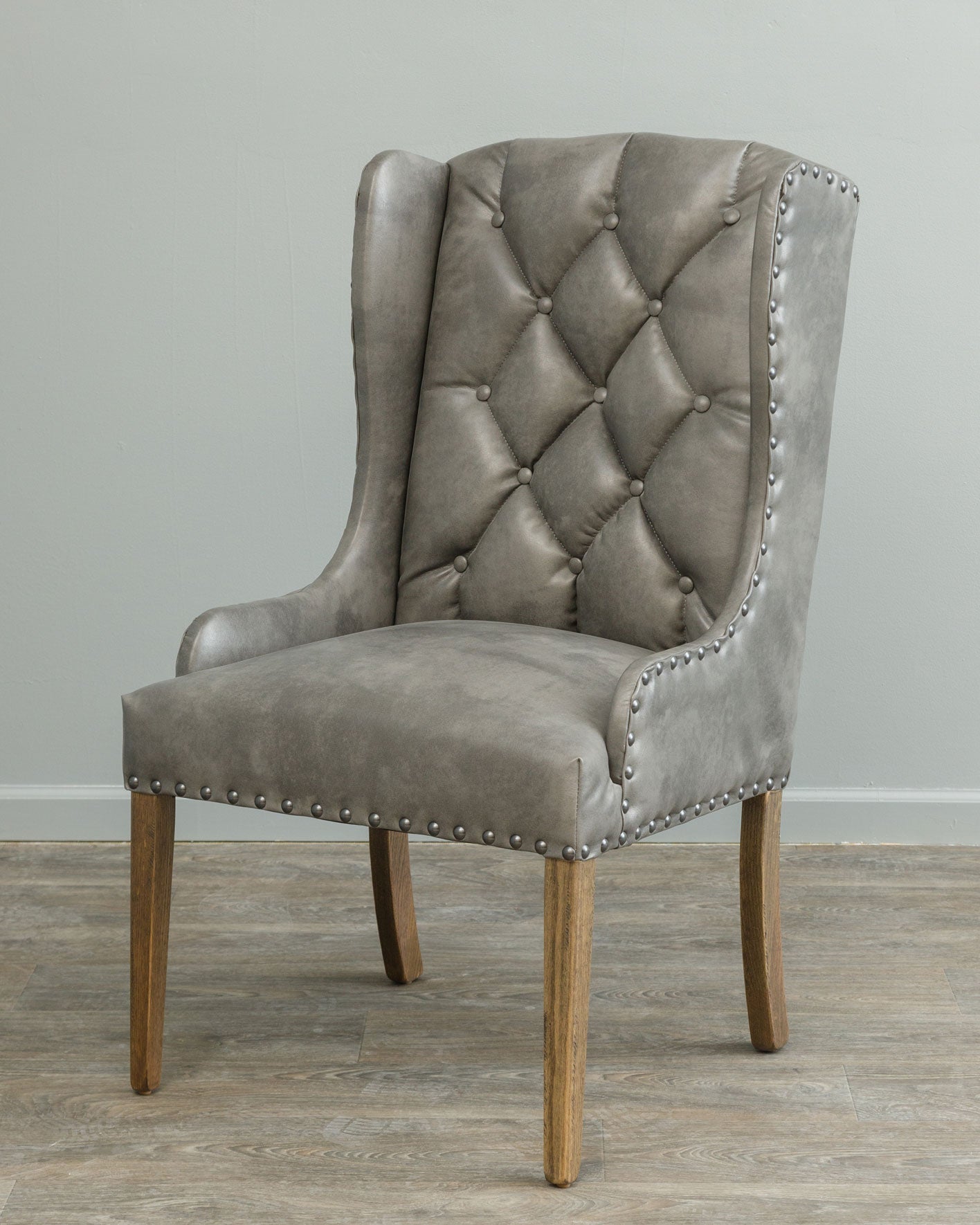 Wingback dining chair upholstered in gray tufted faux leather with pewter nailhead trim and weathered oak legs