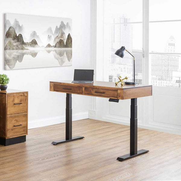 Walnut-toned adjustable standing desk with two drawers on black lift columns, laptop and lamp in a bright city office