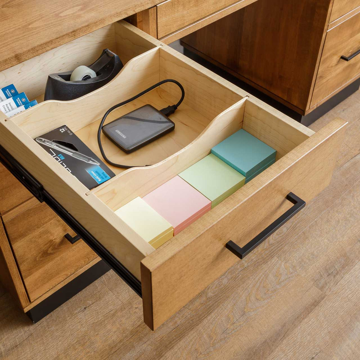 Open desk drawer with maple organizer holding tape dispenser, external drive, pens and colored sticky note pads