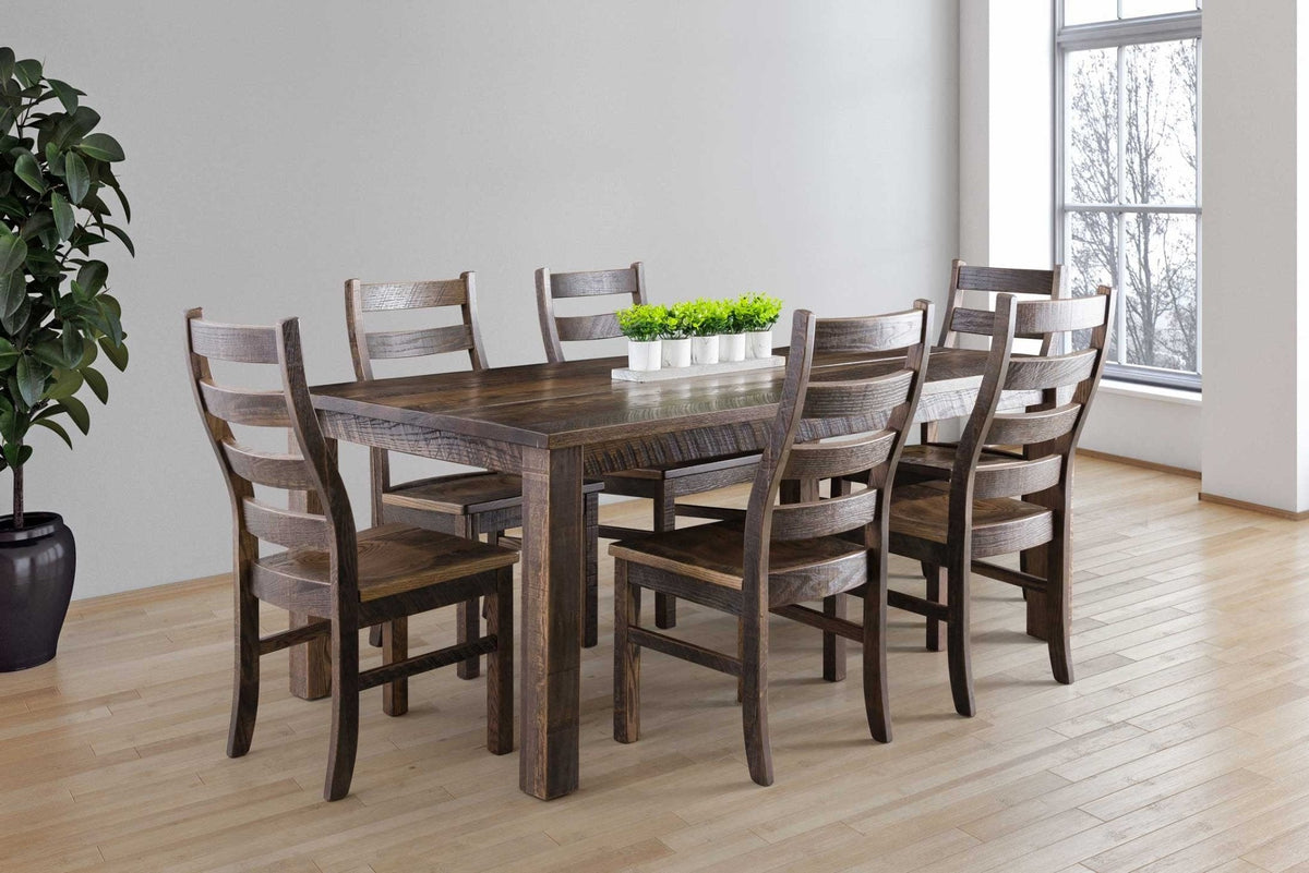 Weathered brown farm dining table with chunky square legs and six ladder back chairs in a bright dining room